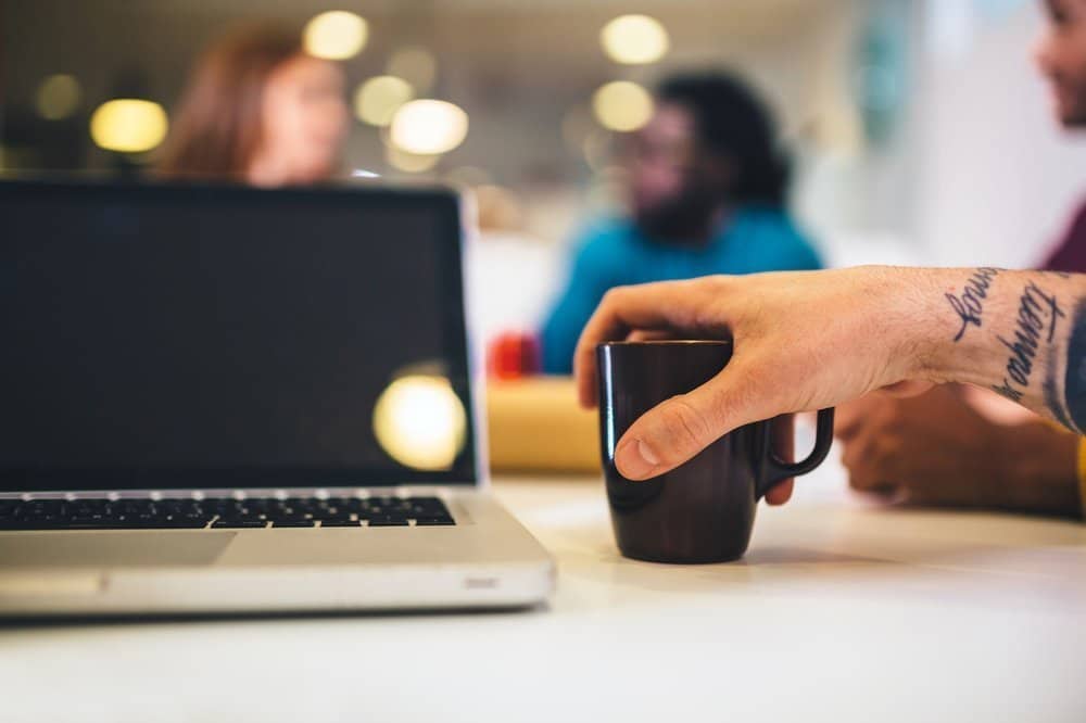 Close up view of incognito man holding mug next to laptop on table.