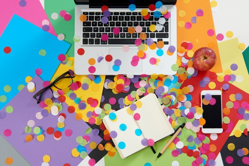 A laptop, glasses, notebook, and a phone on a desk covered with colorful paper and confetti.