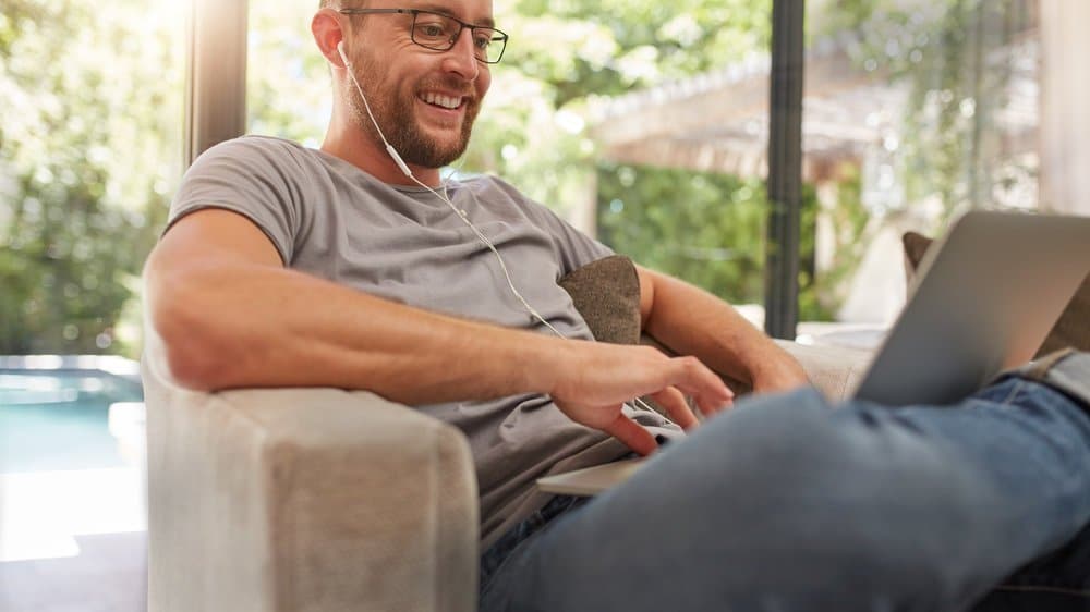 Smiling mature man using his laptop at home, he is sitting on couch with earphones.