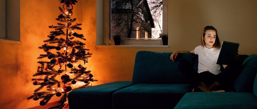 Young girl sitting on sofa in twilight near decorated glowing Christmas tree and using touch pad.