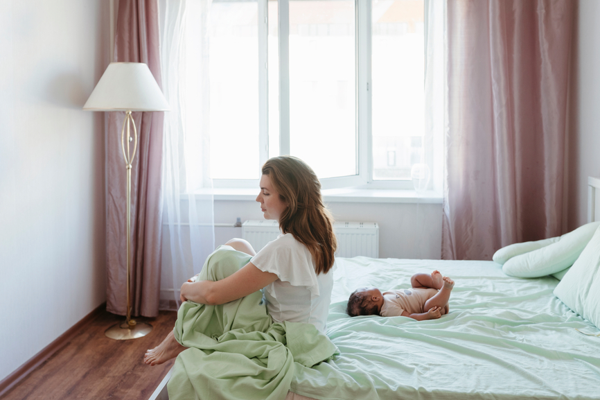A woman looking away while her baby is lying on the bed.