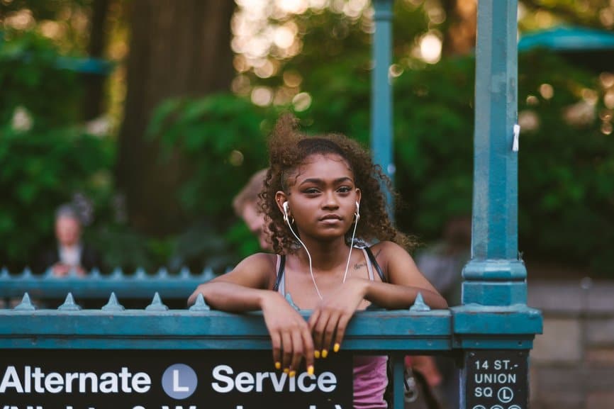 Teenage girl standing by subway station in the city, wearing earbud headphones and looking at camera.