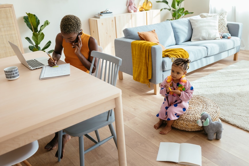 A mother on the phone taking notes while her child plays nearby.