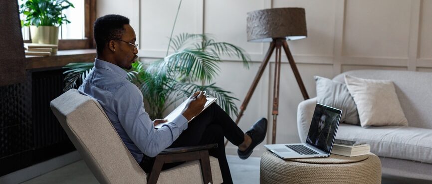 Side view of serious male doctor, using laptop for online therapy session with client and discussing problems with him.