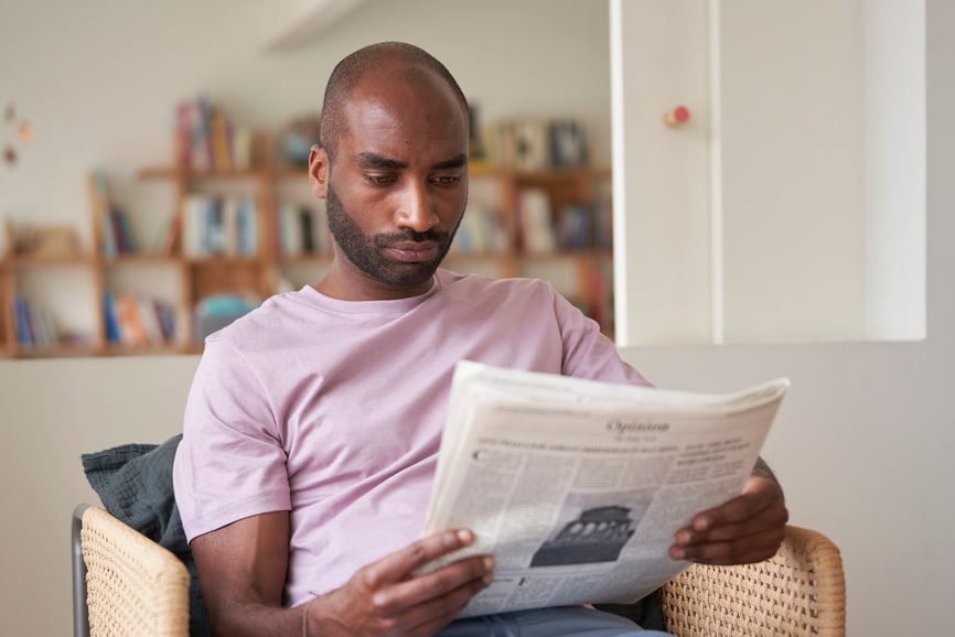 One man with beard in pink t-shirt reading last news in the paper sitting in chair at home.