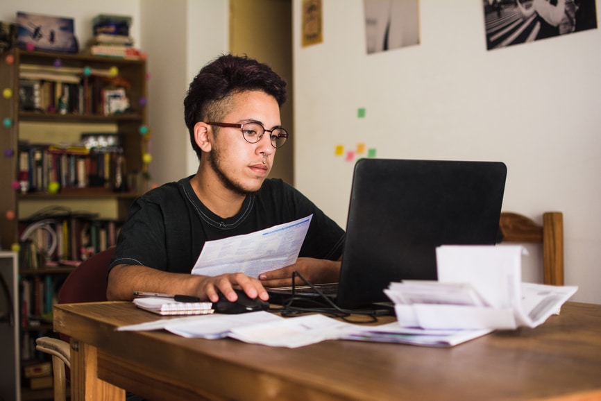 Young man at home working on his personal finances. He is sitting at a wooden table with a black laptop and bills, doing some paper work. He uses glasses and a black t-shirt.