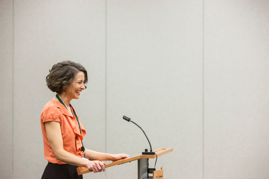 Caucasian businesswoman speaking from a podiium to an audience in a conference room seminar setting.