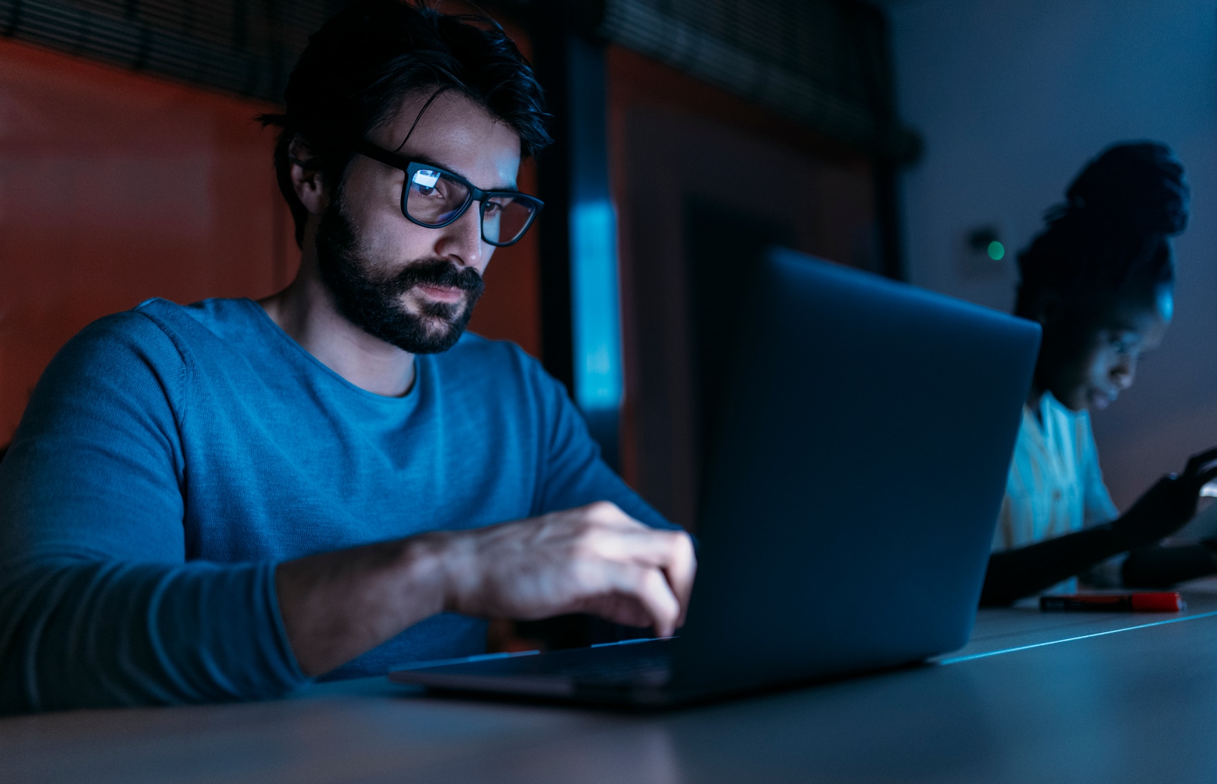 Side view of focused bearded male entrepreneur using netbook while working in dark office with screen hanging on wall near multiethnic coworkers
