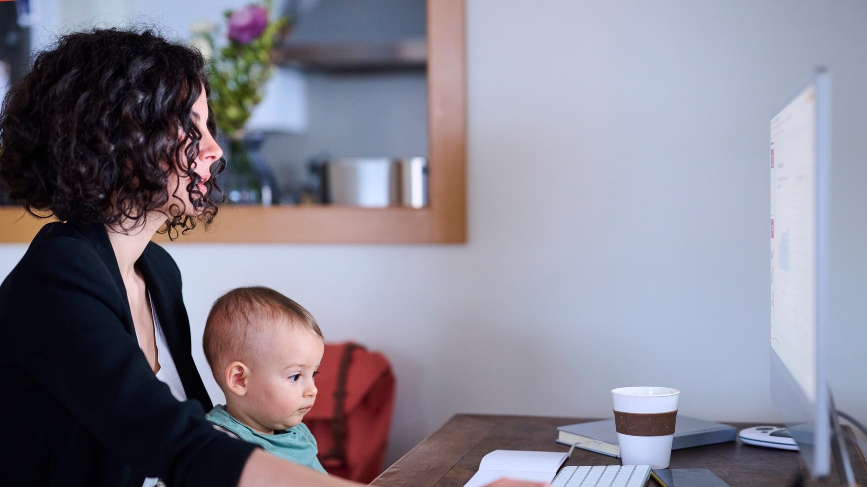 Side view of businesswoman with sleepy toddler sitting at desk and browsing data on computer while working on freelance project in obscure home office in evening.