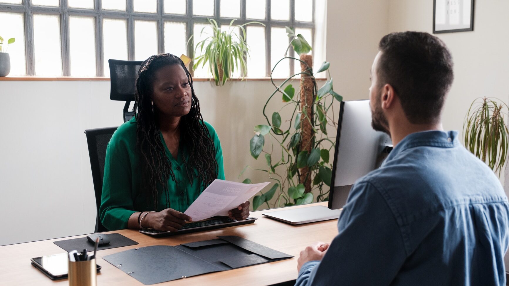 Concentrated female boss interviewing an anonymous man, a job candidate, sitting at her desk at office. She is holding a document, while listening and looking at him.