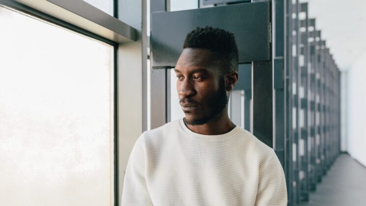 Young black man wearing white sweater with pensive expression looking out window