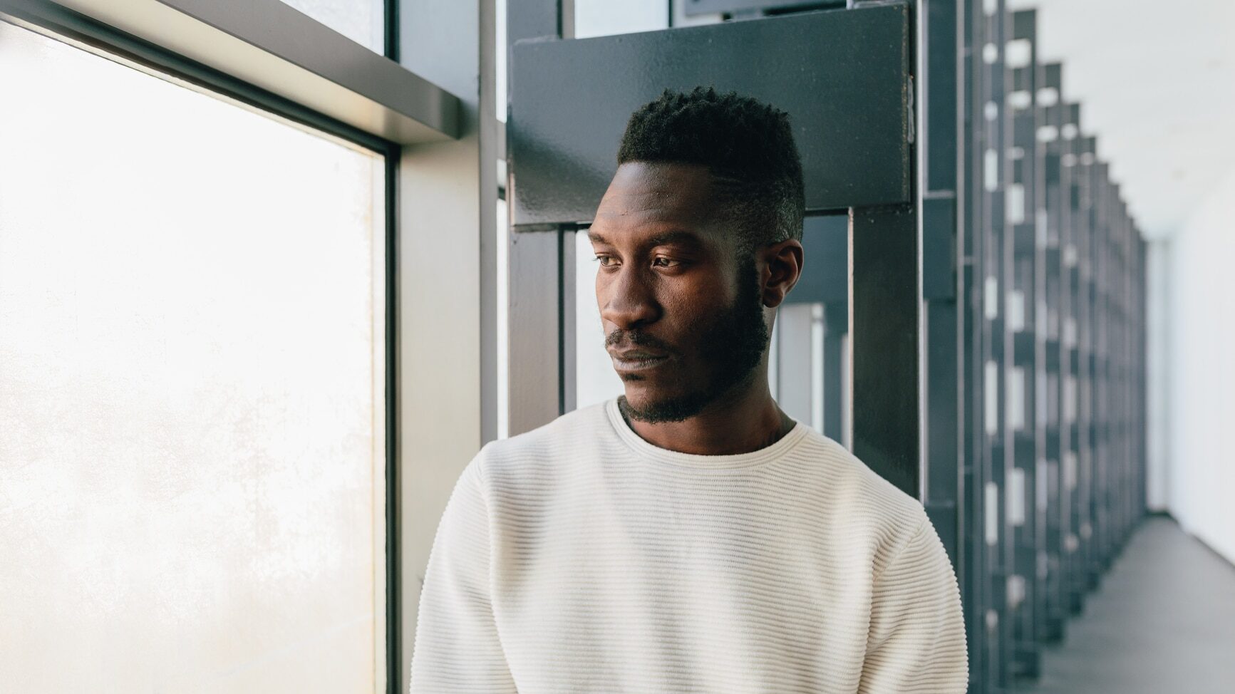 Young black man wearing white sweater with pensive expression looking out window