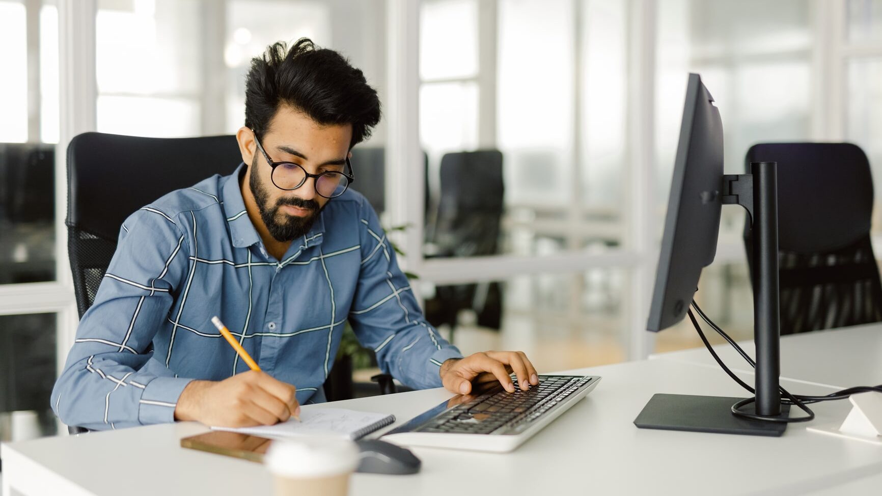 Concentration entrepreneur using PC technology office. Focused employee in glasses seating at modern workplace with computer and cup on it, holding pen and jotting down important details