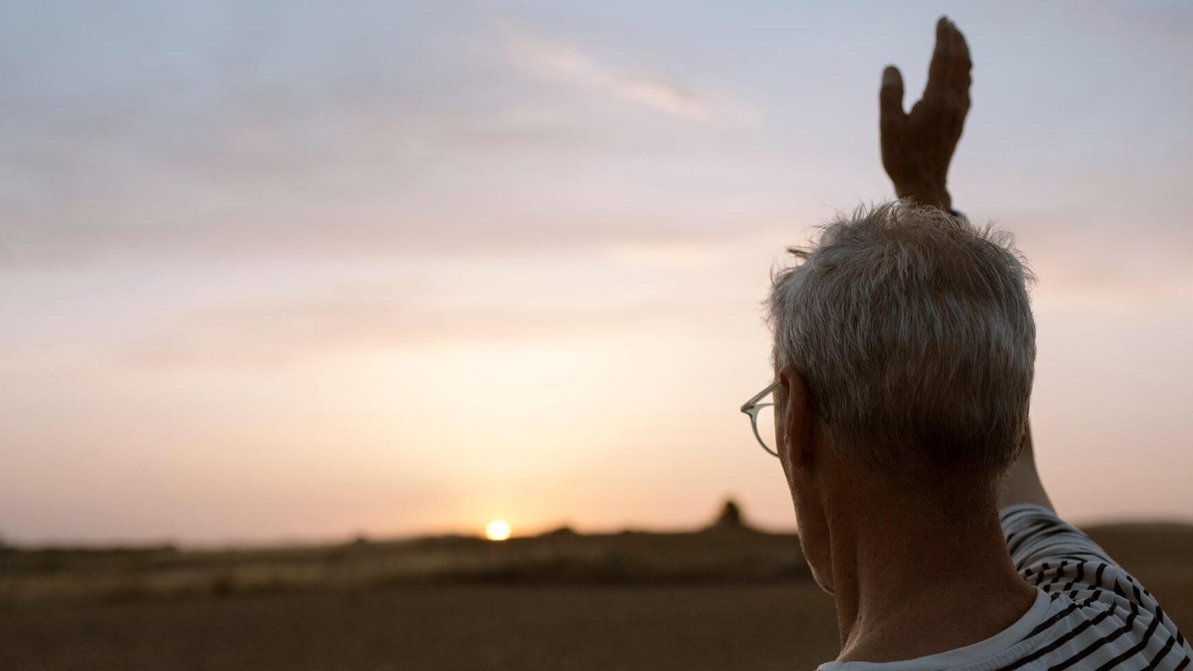 Senior grey-haired man wearing a striped t-shirt and using glasses waving at sun at sunset in the countryside
