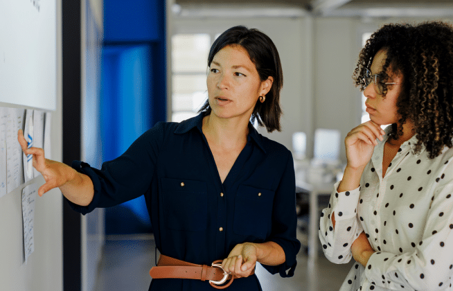 Two women stand by a whiteboard. One woman points at notes on the board while explaining something; the other listens attentively with a thoughtful expression, hand on her chin. Both are in an office setting.