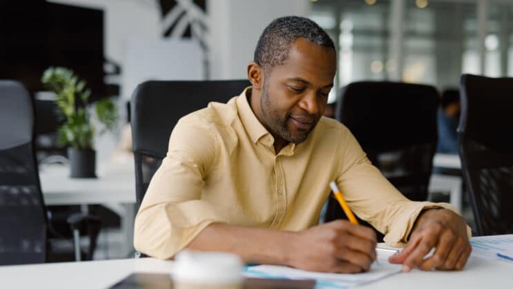 Businessperson paperwork note workplace data. Black man in good spirit, getting right on files and marking on them with pencil in empty workroom with gadget and disposable cup near him