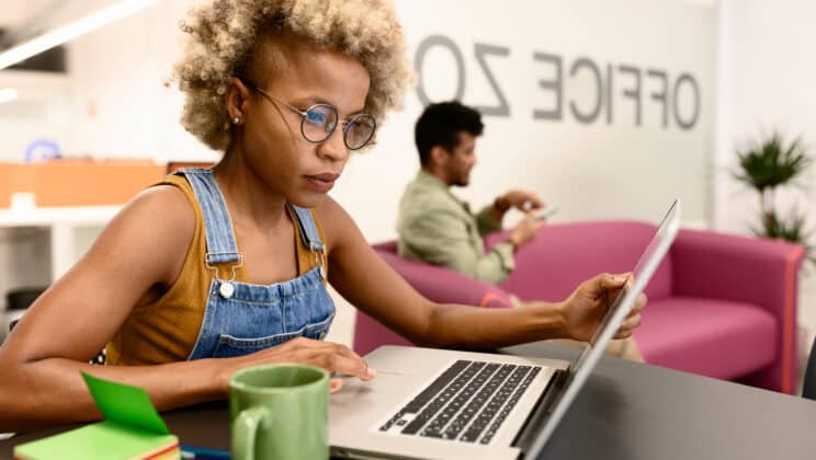 One south american employee with dyed blonde hair in eyeglasses wearing top and denim overall browsing laptop