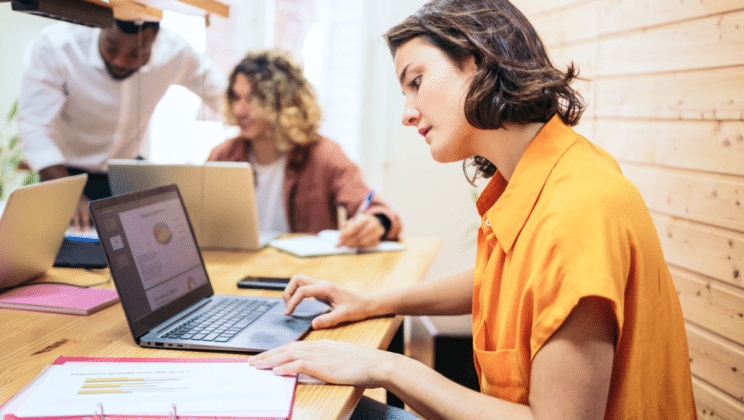 Three people work at a table with laptops and documents. The focus is on a woman in an orange shirt reviewing a report on her laptop, while two colleagues collaborate in the background.
