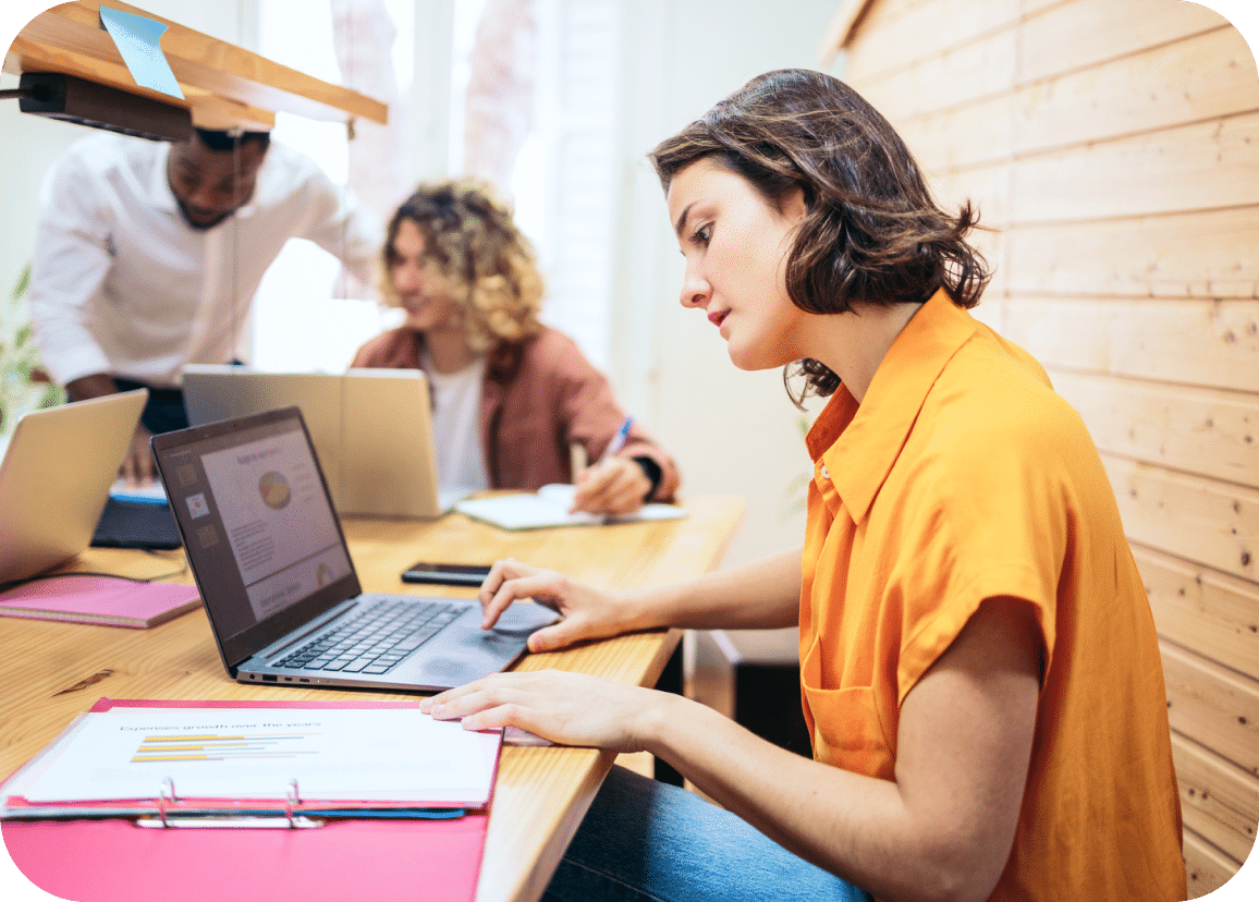 Three people work at a table with laptops and documents. The focus is on a woman in an orange shirt reviewing a report on her laptop, while two colleagues collaborate in the background.