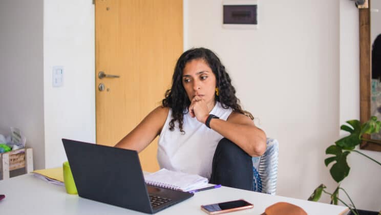 Woman sitting comfortably at home desk, using her laptop looking at the screen with a reluctant, serious, bore expression.