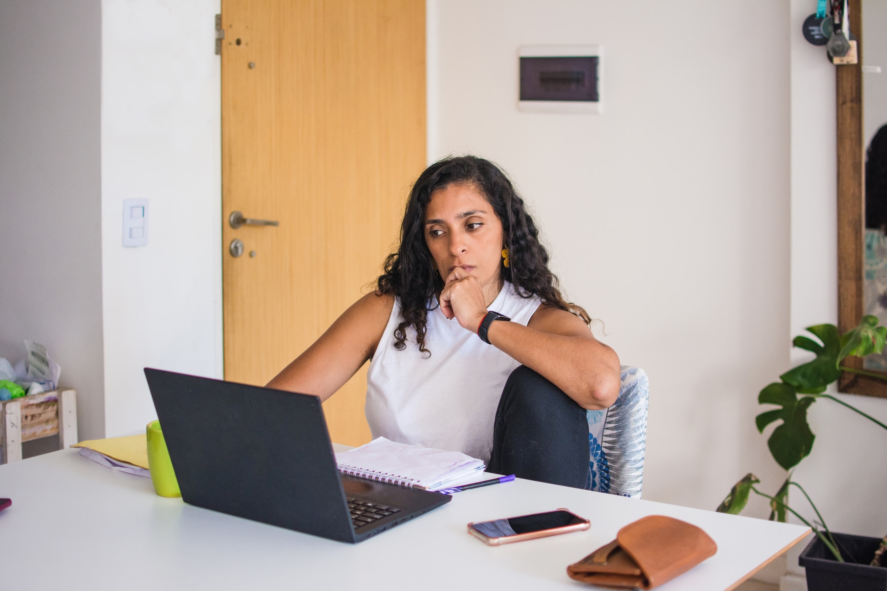 Woman sitting comfortably at home desk, using her laptop looking at the screen with a reluctant, serious, bore expression.