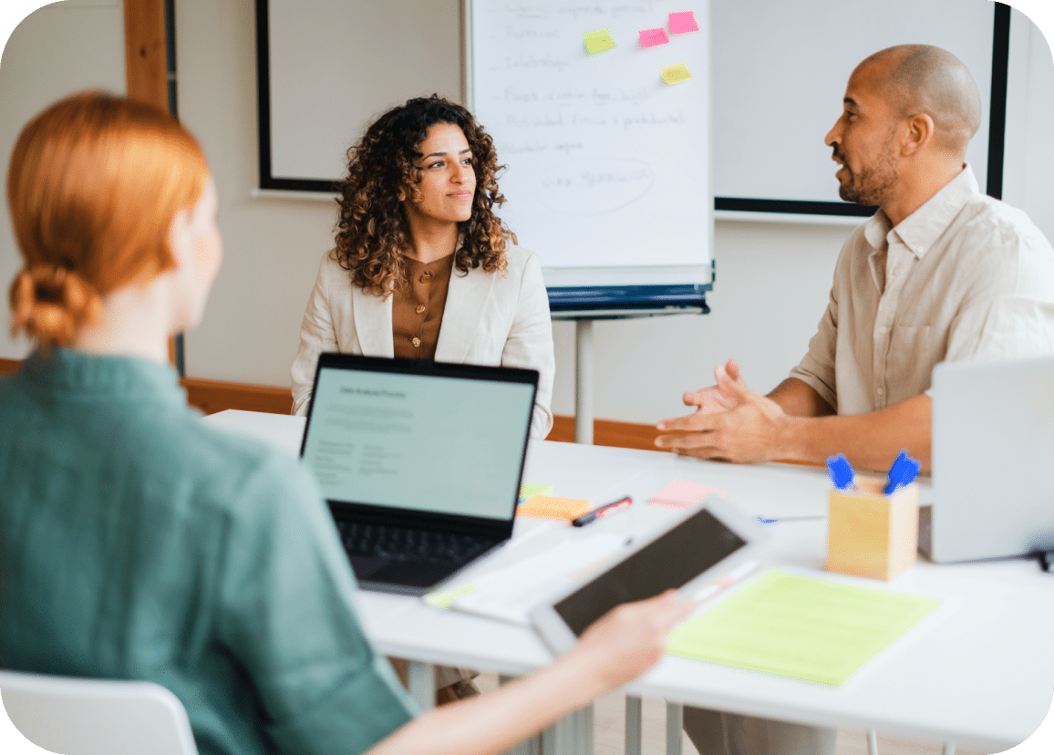 Three people sit around a table having a discussion, with laptops, notebooks, and sticky notes visible. A whiteboard with colorful sticky notes is in the background.