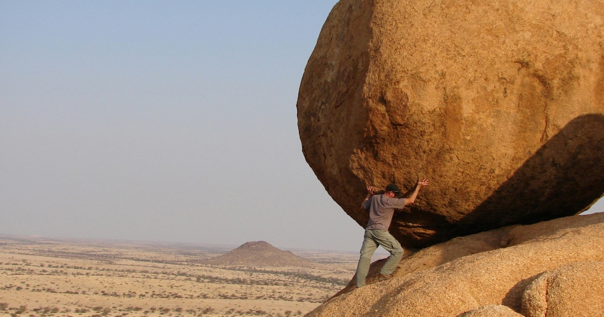 A person pushes against a giant boulder on a rocky hill with a vast desert landscape and distant hills in the background.