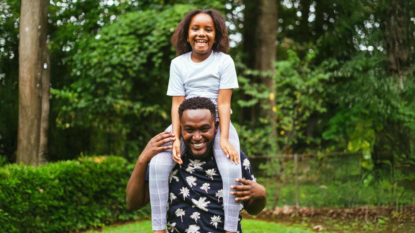A smiling man gives a young girl a piggyback ride on his shoulders in a lush green outdoor setting, both appearing happy and playful.