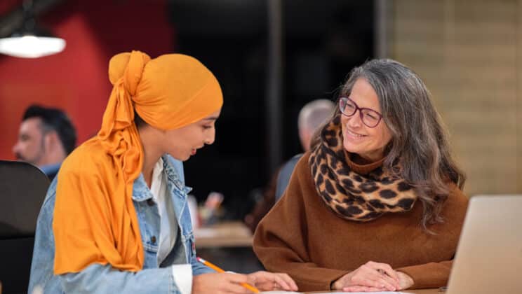 One worker in yellow hijab and denim jacket holding a pencil explaining her report to a cheerful CEO with long gray hair in stylish scarf and brown sweater wearing eyeglasses. Women business meeting together concept