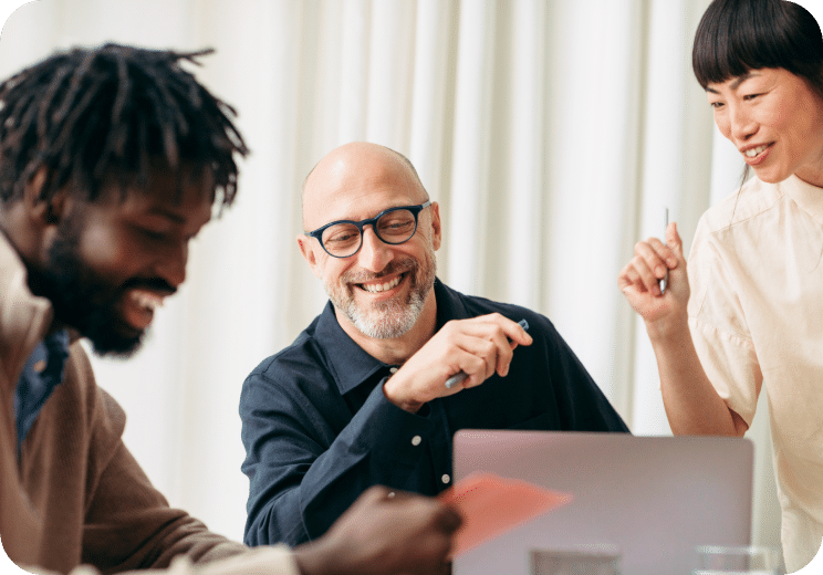 Three people sit at a table, smiling and discussing something while looking at a laptop. One person holds a pen, and the atmosphere appears collaborative and friendly.
