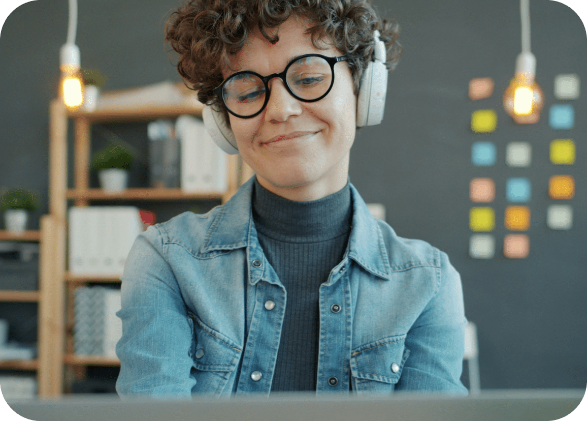 A person with curly hair and glasses, wearing headphones and a denim jacket, smiles while working at a desk. Behind them are shelves, hanging lights, and a wall with colorful sticky notes.