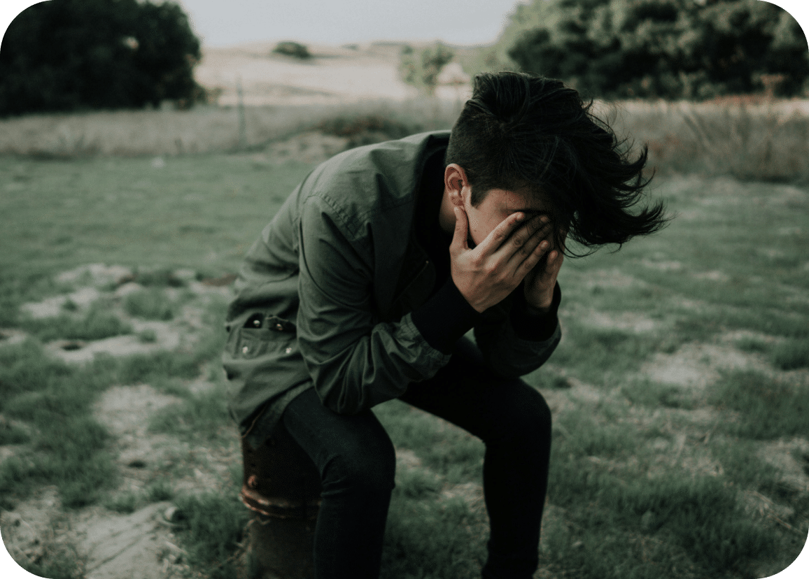 A person sits on a stool outdoors in a grassy field, wearing a green jacket with hands covering their face, appearing distressed or upset. Trees and hills are visible in the background under a cloudy sky.