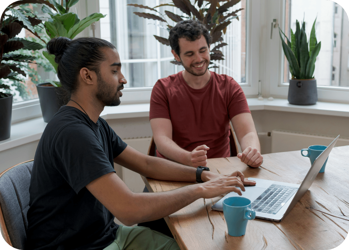 Two people sit at a wooden table with a laptop, smiling and talking. There are blue mugs and potted plants on the table and windowsill behind them.