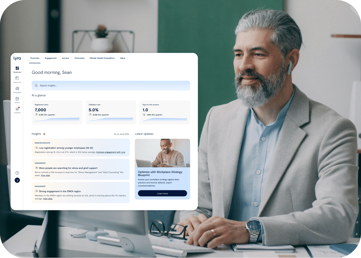 A man with gray hair and beard, wearing a gray jacket, sits at a desk with a laptop. On the left, a computer screen displays a dashboard with insights and metrics.