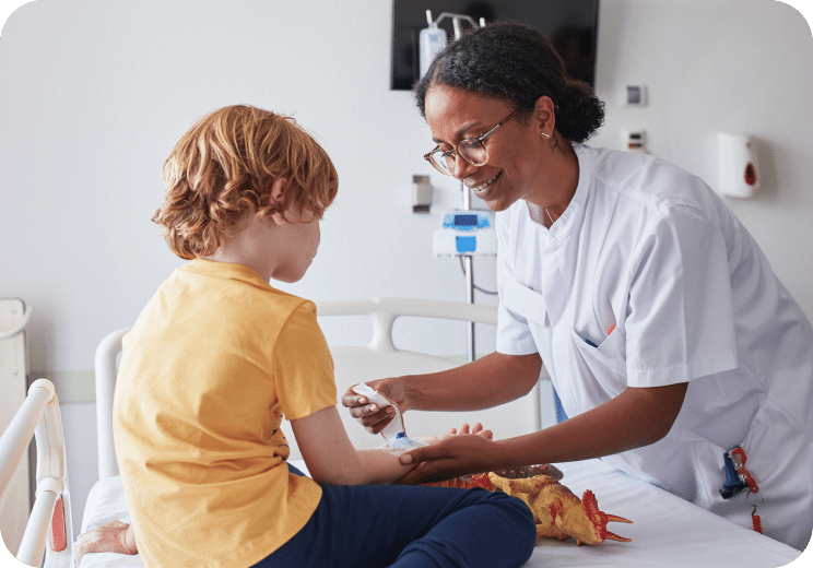 A nurse smiles while preparing to give an injection to a young child sitting on a hospital bed, with a toy dinosaur nearby.