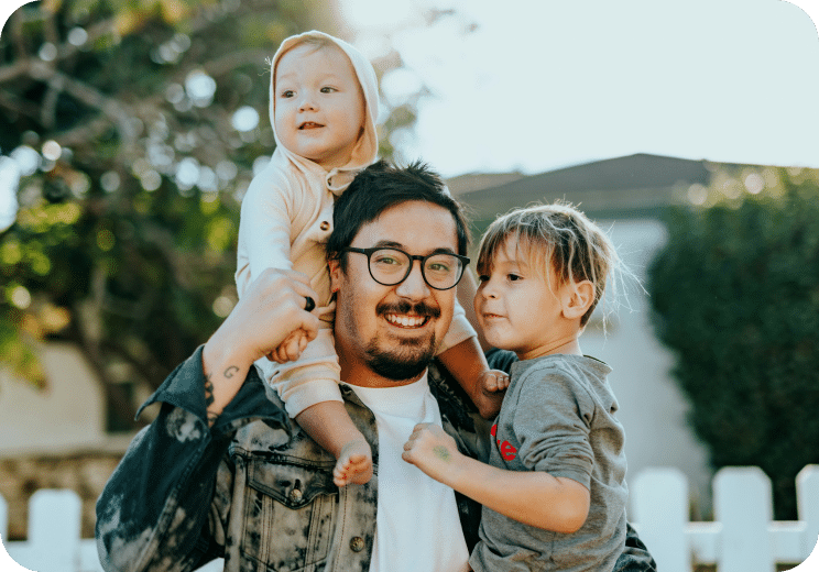 A smiling man with glasses holds a young child on his shoulders while another child stands close to him, all outdoors on a sunny day with greenery and a white fence in the background.