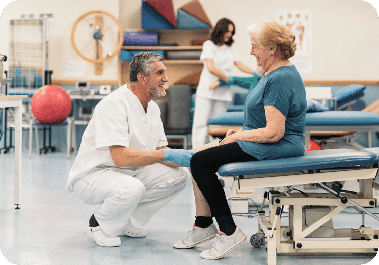 A healthcare worker in white scrubs and gloves kneels, smiling and talking with an older woman sitting on a treatment table in a physical therapy room. Another person works in the background.