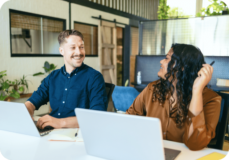 Two coworkers sit at a desk with laptops, smiling and looking at each other in a modern office space with plants and glass dividers in the background.