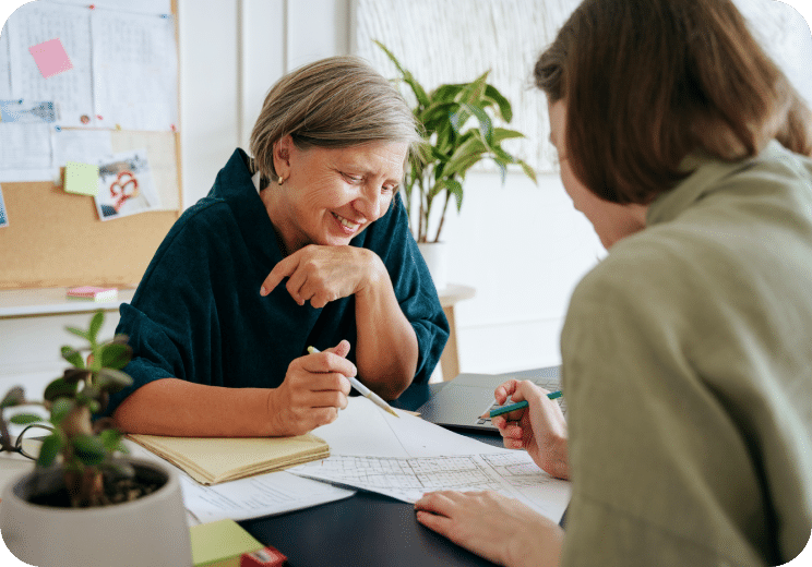 Two people sit at a desk, smiling and discussing papers and charts. One person points at a document while the other holds a pen. A plant and notebook are on the table; a bulletin board is in the background.