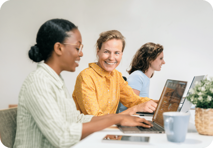 Three women sit at a table with laptops, working and smiling. One woman in a yellow shirt looks at the camera, while the others focus on their screens. A mug and potted plant are on the table.