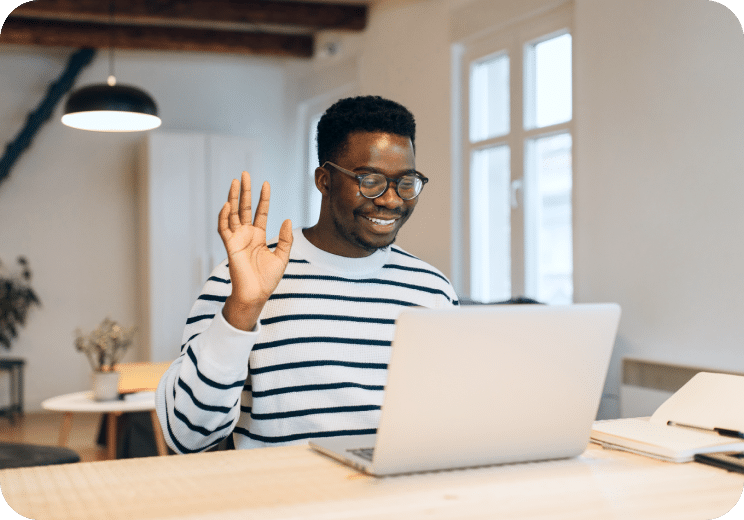Smiling person wearing glasses and a striped sweater sits at a desk, waving at a laptop during a video call. Bright, modern room with large windows and wooden beams in the background.