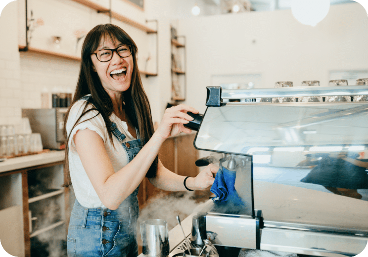 A smiling barista wearing glasses and overalls stands behind a coffee machine in a bright café, appearing cheerful while preparing coffee.