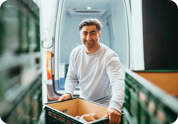 A man smiling while lifting a box from the back of a van, surrounded by green crates, possibly making a delivery.