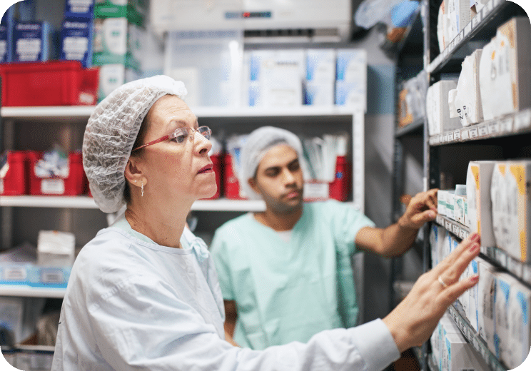 Two medical staff wearing hairnets and scrubs organize and check boxes of supplies on shelves in a storage room.