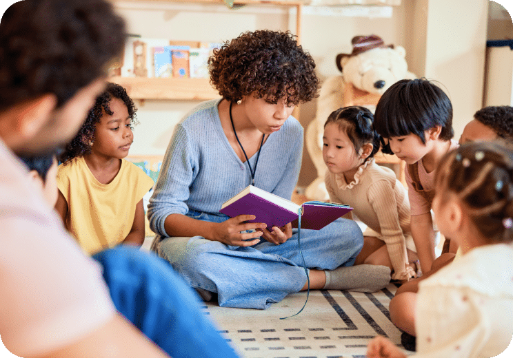 A teacher sits on the floor reading a book to a group of young children who are listening closely, surrounded by bookshelves and toys in a classroom setting.