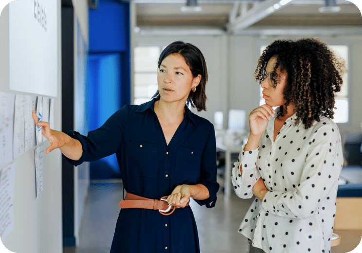 Two women stand in an office, one pointing at documents on a wall while the other listens and observes thoughtfully.