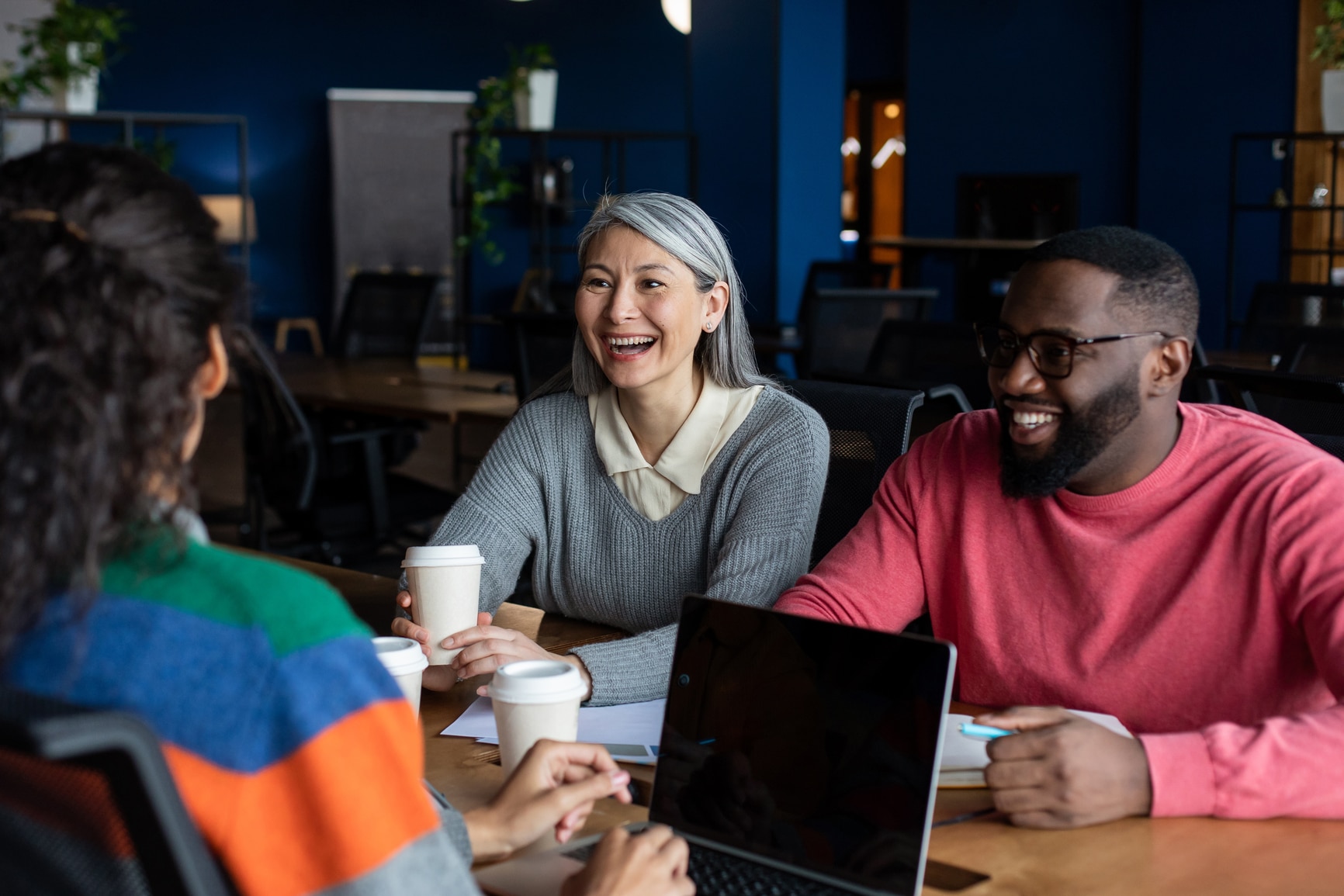 Three people sit at a table with coffee cups, smiling and talking in a modern office setting. One person has gray hair, another wears glasses and a pink sweater, and the third has curly hair and a colorful sweater.