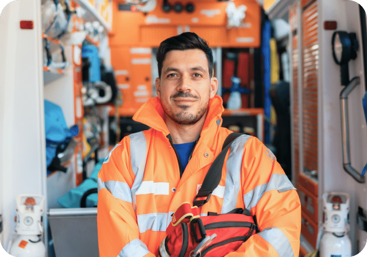 A man in an orange reflective jacket stands inside an ambulance, holding a red medical bag and smiling confidently at the camera.
