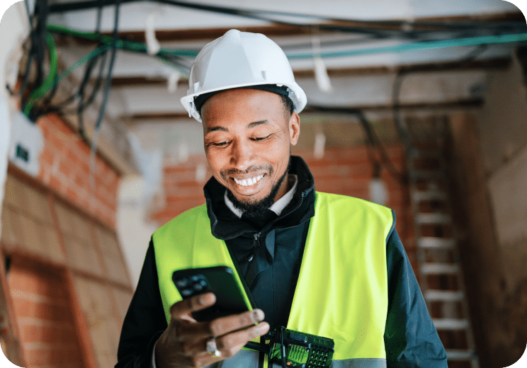A construction worker wearing a white hard hat and yellow safety vest smiles while looking at his phone inside a building under construction.