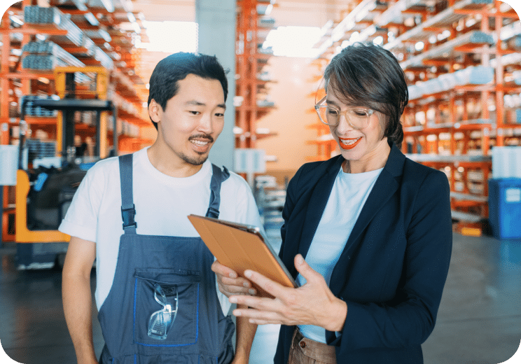 A man in overalls and a woman in business attire look at a tablet together while standing in a warehouse filled with shelves and boxes.